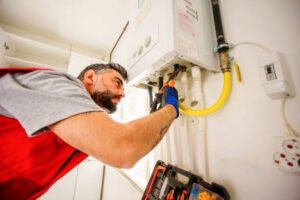 A man wearing a red shirt and vest repairs a water heater in a home setting, focused on his task, related to electrical and plumbing troubleshooting.