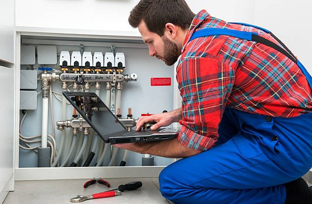 A man in overalls focused on working on a laptop at a desk, surrounded by tools and equipment, related to electrical and plumbing troubleshooting.