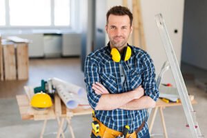 A man wearing a plaid shirt and hard hat stands confidently in front of a construction site, overseeing the work, representing experienced handyman services.