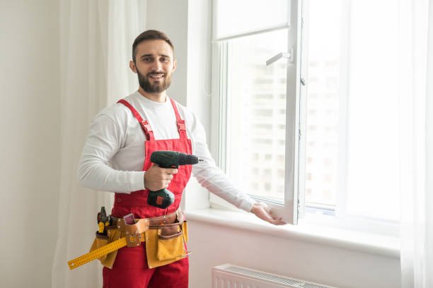 A man in blue overalls stands holding a drill in one hand and a hammer in the other, ready for a construction task, representing experienced handyman services.