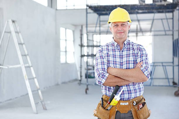A man wearing a hard hat stands confidently in a construction area, surrounded by building materials and equipment, representing a local handyman.