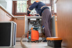 A man repairs a kitchen sink while using a vacuum to clean up debris, demonstrating plumbing troubleshooting skills.