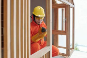 A man in an orange safety suit providing handyman services for door and window framing, repairing a wooden house amidst construction materials in Huntington Beach.