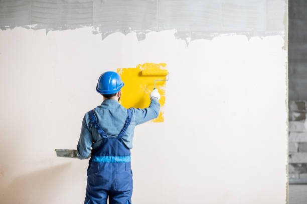 A painting contractor in Tucson, AZ wearing blue overalls and carefully applying bright yellow paint to a wall